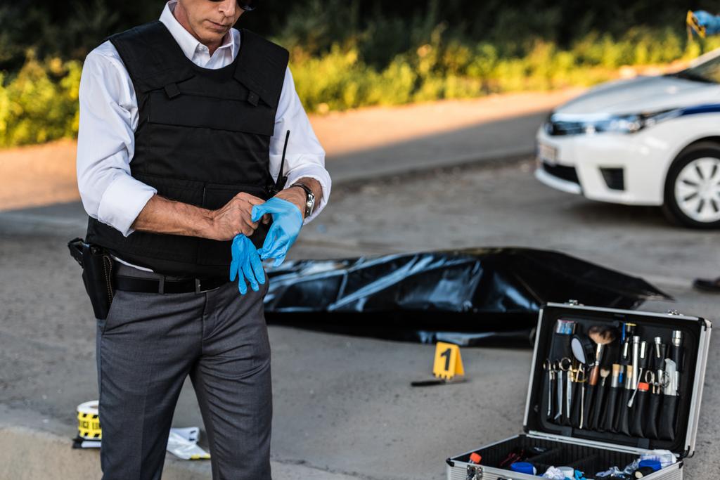 visão parcial do policial com arma no coldre colocando luvas de látex na cena do crime com cadáver no saco do corpo e caso com ferramentas de investigação
  - Foto, Imagem