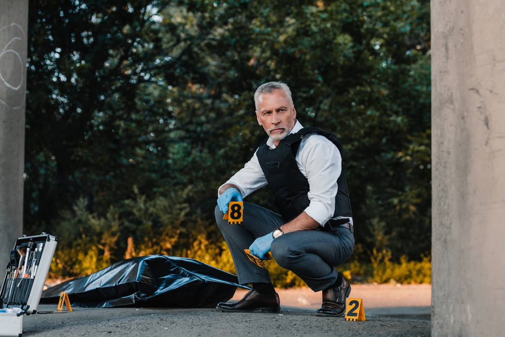 ernster männlicher Polizeibeamter in Latexhandschuhen sitzt am Tatort mit Leiche im Leichensack  - Foto, Bild