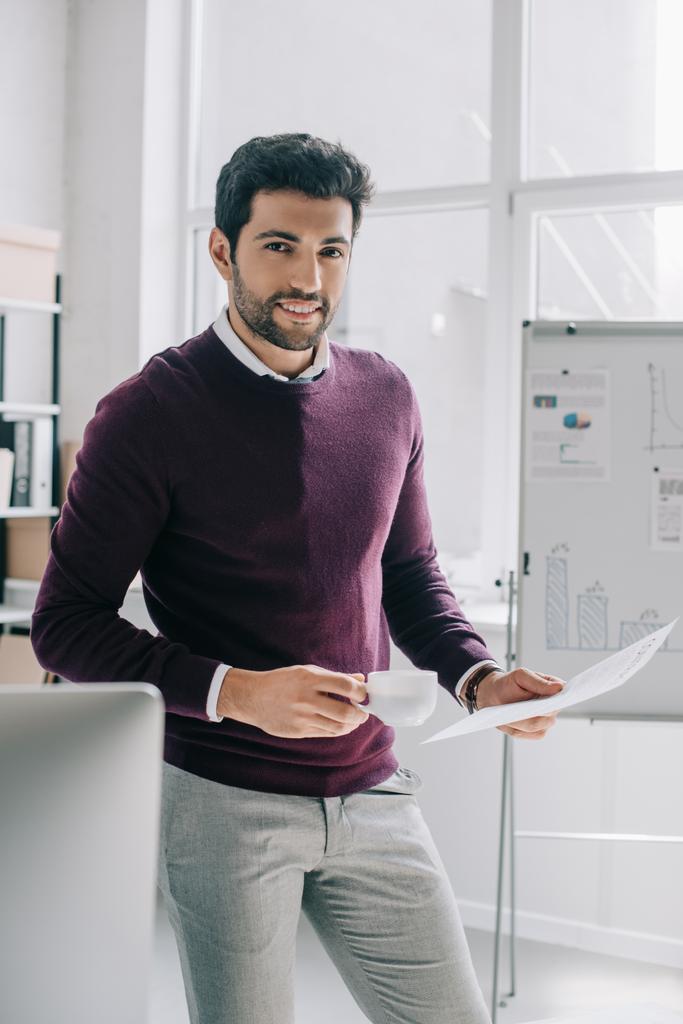 красивый бизнесмен в бордовом свитере с чашкой кофе и документами в офисе
 - Фото, изображение