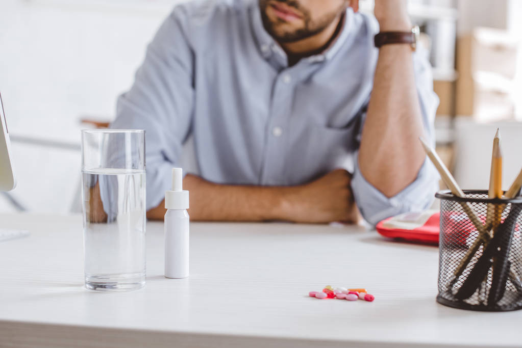 обрезанный образ больного менеджера, опирающегося на стол с лекарствами в офисе
 - Фото, изображение