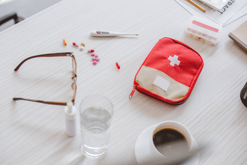 high angle view of first aid kit, glass of water, cup of coffee and pills on table in office - Photo, Image