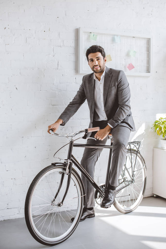 smiling businessman sitting on bicycle in office and looking at camera - Photo, Image