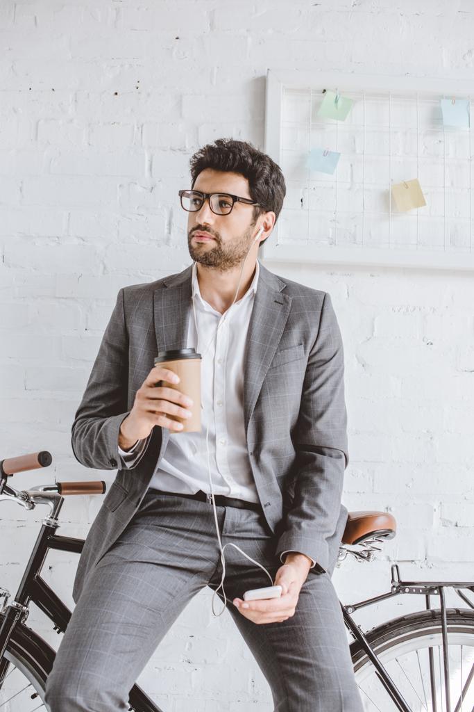 businessman listening music with smartphone, holding cup of coffee and leaning on bike in office - Photo, Image