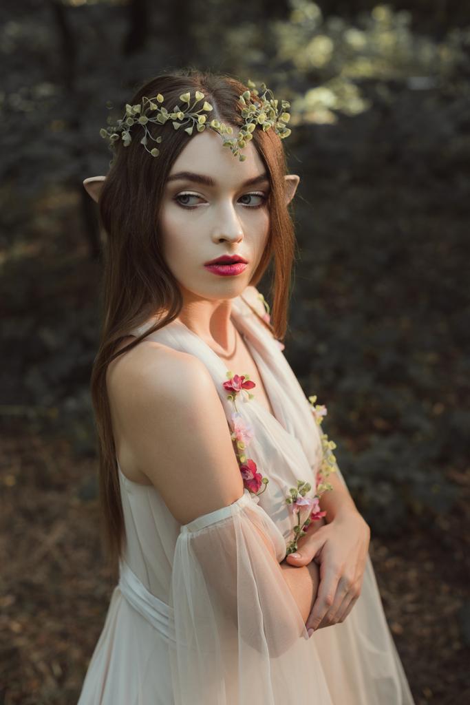 kaunis nainen tonttu kukka mekko ja seppele seisoo ristissä kädet metsässä
 - Valokuva, kuva