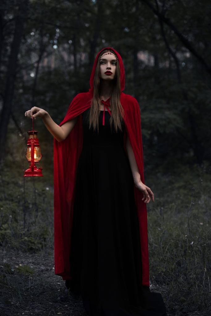 joven mujer mística en capa roja con lámpara de queroseno caminando en bosques oscuros
  - Foto, imagen