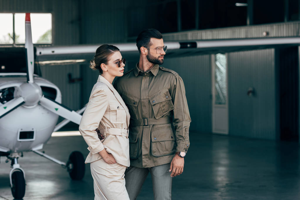pareja joven de moda en chaquetas mirando lejos cerca de avión en hangar
  - Foto, Imagen