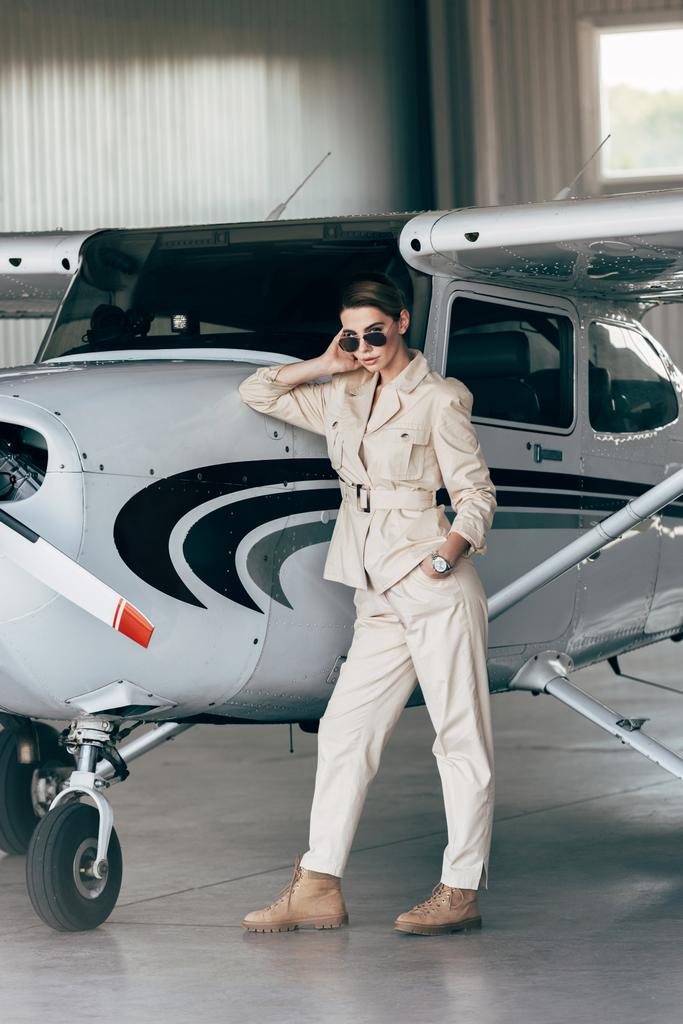 fashionable young woman in sunglasses and jacket posing near aircraft in hangar - Photo, Image