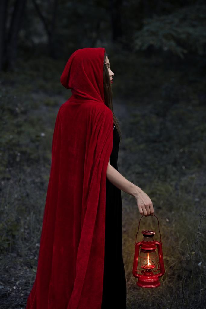 μυστικιστική κορίτσι με κόκκινο μανδύα περπάτημα σε σκοτεινό δάσος με λάμπες κηροζίνης - Φωτογραφία, εικόνα