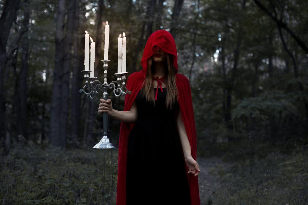 menina mística em manto vermelho e capuz segurando candelabro com velas na floresta escura
 - Foto, Imagem