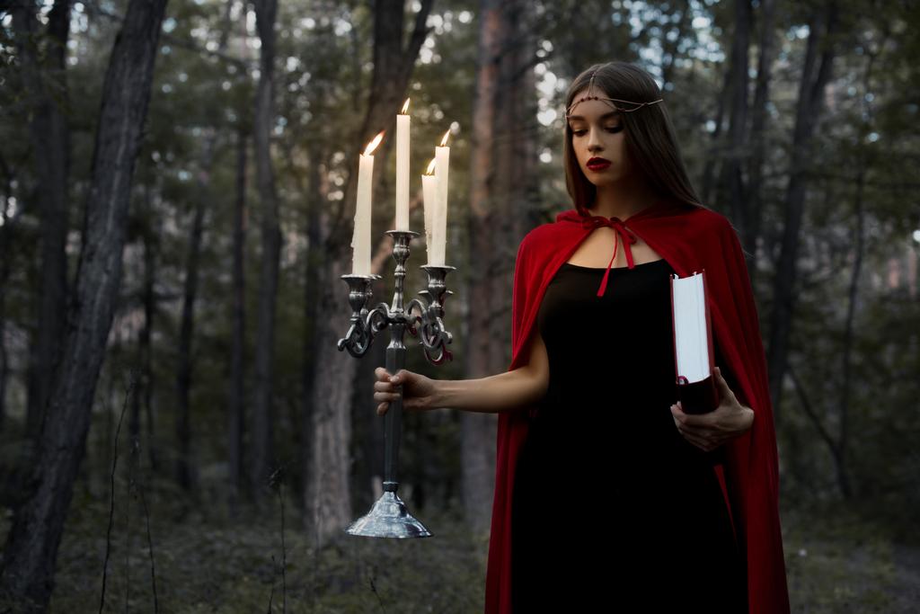 chica mística en capa roja sosteniendo candelabro con velas encendidas y libro mágico en bosques oscuros
 - Foto, imagen