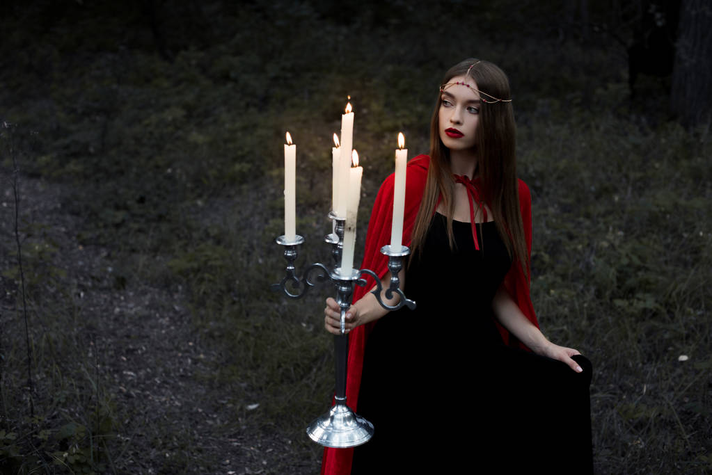 elegantní mystic dívka v červený plášť, který drží svícen s hořící svíčky v temném lese - Fotografie, Obrázek