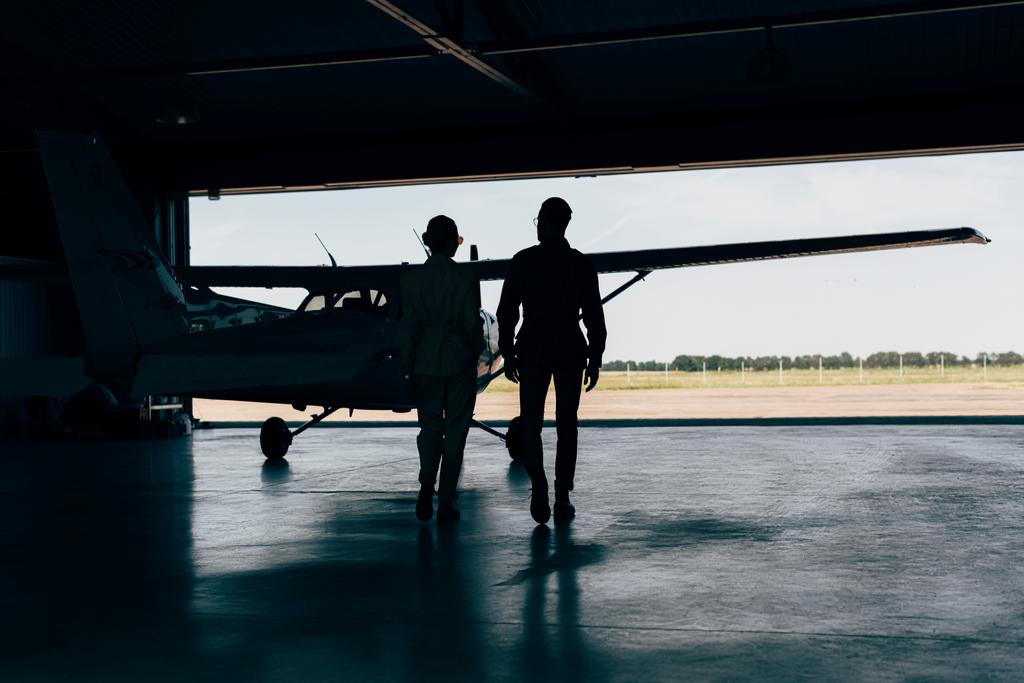Achteraanzicht van de silhouetten van stijlvolle paar wandelen in de buurt van vliegtuig in loods  - Foto, afbeelding