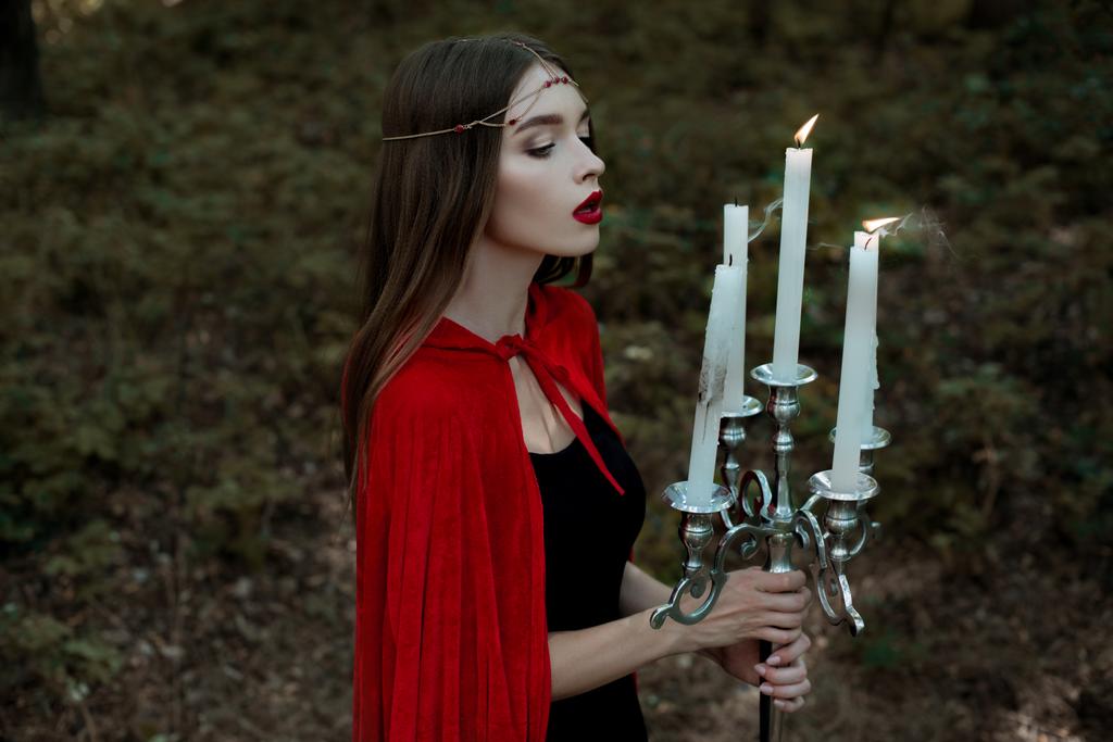 menina mística elegante em manto vermelho e grinalda soprando velas em candelabro na floresta escura
 - Foto, Imagem