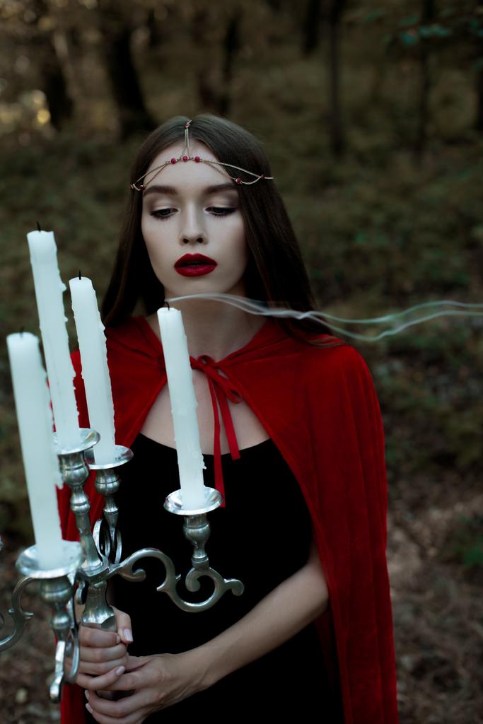 attractive mystic girl in red cloak blowing candles in forest - Photo, Image