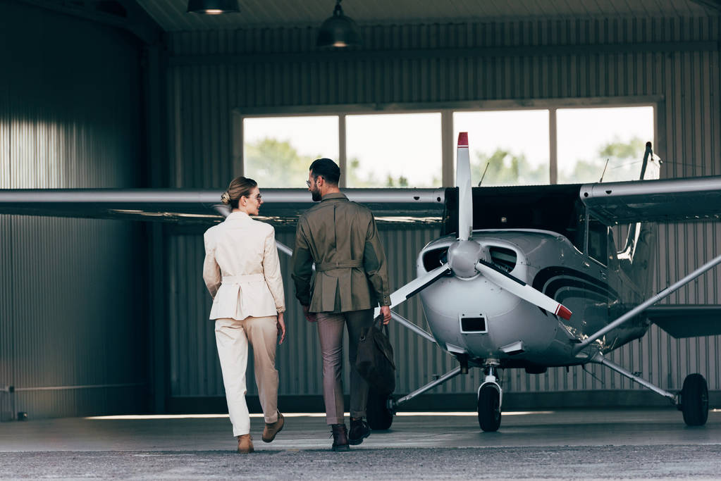 vista trasera del hombre que lleva el bolso y caminar con novia con estilo cerca del hangar con el avión
  - Foto, imagen
