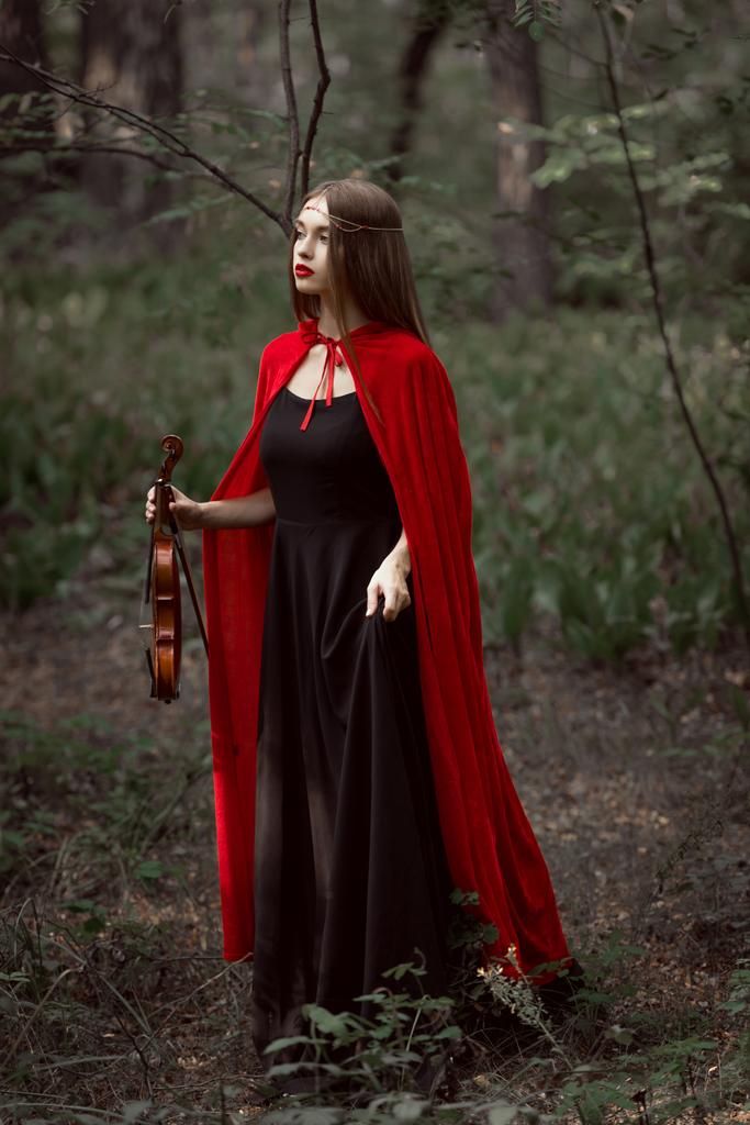 aantrekkelijke mystic vrouw in rode mantel holding viool in donker bos - Foto, afbeelding