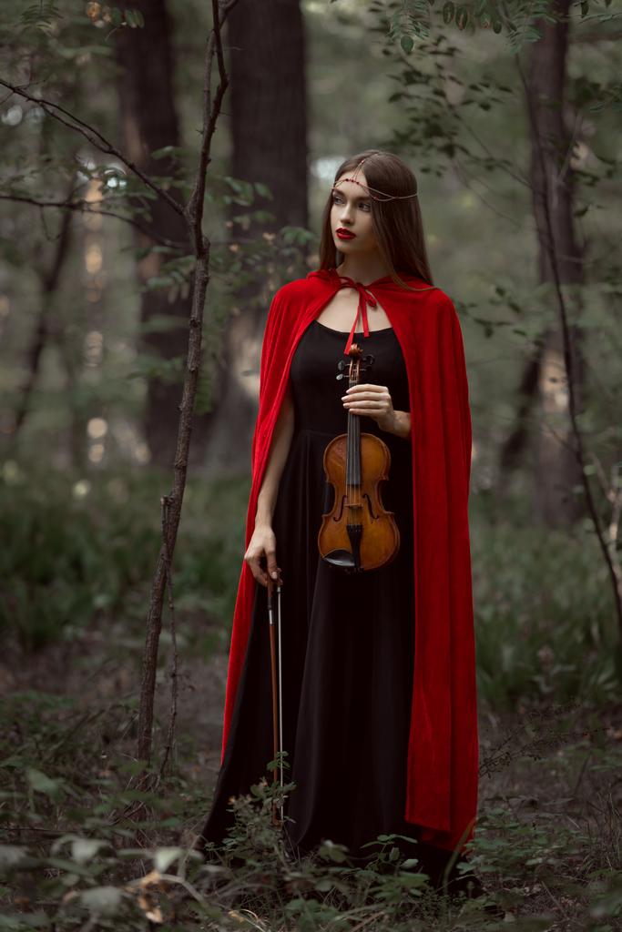 κομψό όμορφη γυναίκα στο μαύρο φόρεμα και κόκκινο μανδύα, κρατώντας βιολί στο δάσος - Φωτογραφία, εικόνα