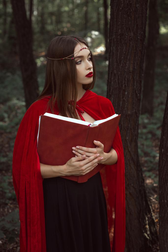 beautiful mystic girl in red cloak and wreath holding magic book in forest - Photo, Image
