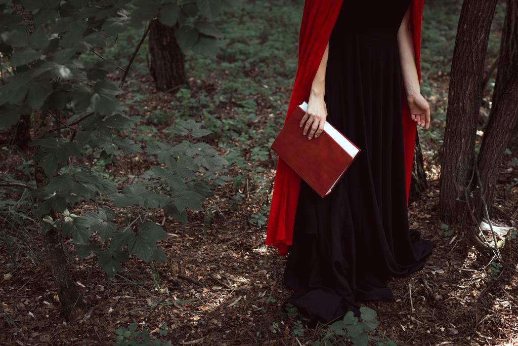 περικοπεί δείτε κομψό κορίτσι με κόκκινο μανδύα με μαγικό βιβλίο στο δάσος - Φωτογραφία, εικόνα