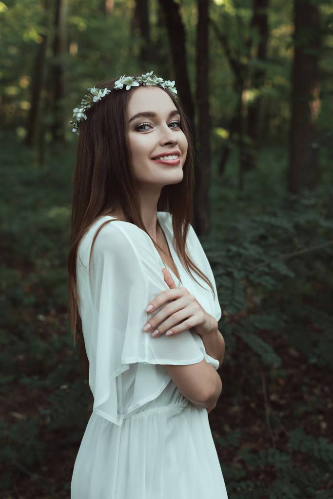attraktives lächelndes Mädchen posiert in elegantem Kleid und Blumenkranz im Wald - Foto, Bild