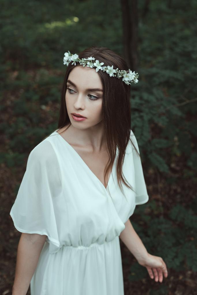 приваблива дівчина позує в білій сукні і квітковому вінку в лісі
 - Фото, зображення