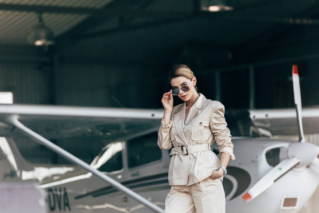 ελκυστική νεαρή γυναίκα σε γυαλιά ηλίου και τζάκετ ποζάρει κοντά αεροσκάφη  - Φωτογραφία, εικόνα