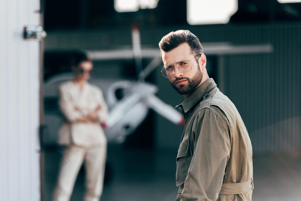 retrato de hombre guapo en gafas y chaqueta mirando a la cámara mientras su novia de pie en el hangar con avión
 - Foto, Imagen