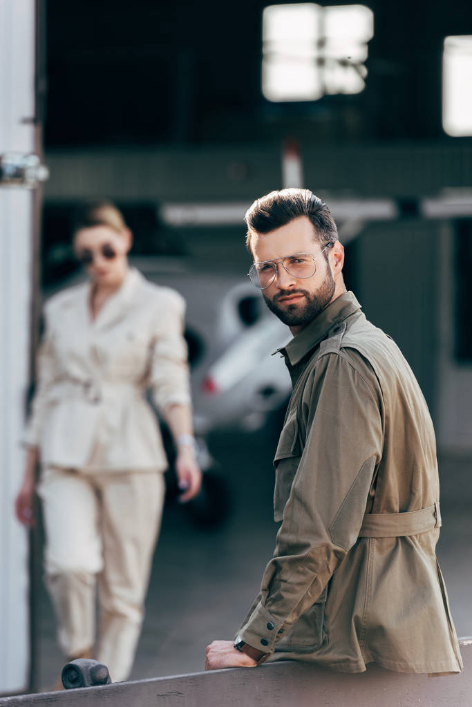 foco seletivo do homem elegante em óculos e casaco olhando para a câmera enquanto sua namorada andando perto do hangar com avião
 - Foto, Imagem
