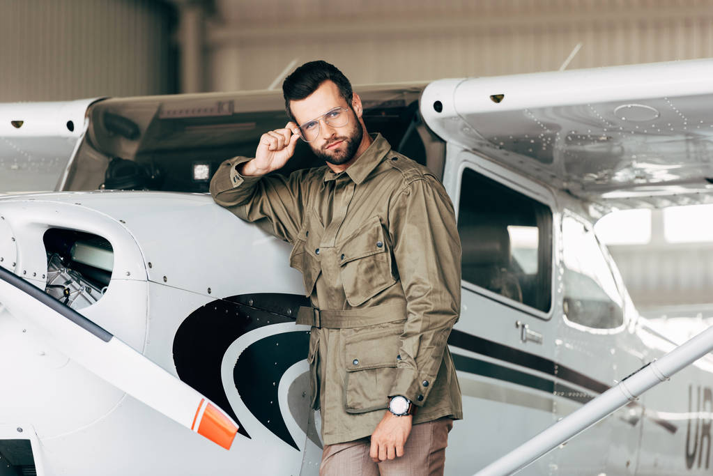 Stilvoller junger Mann in grüner Jacke und Brille blickt in Flugzeugnähe in die Kamera  - Foto, Bild