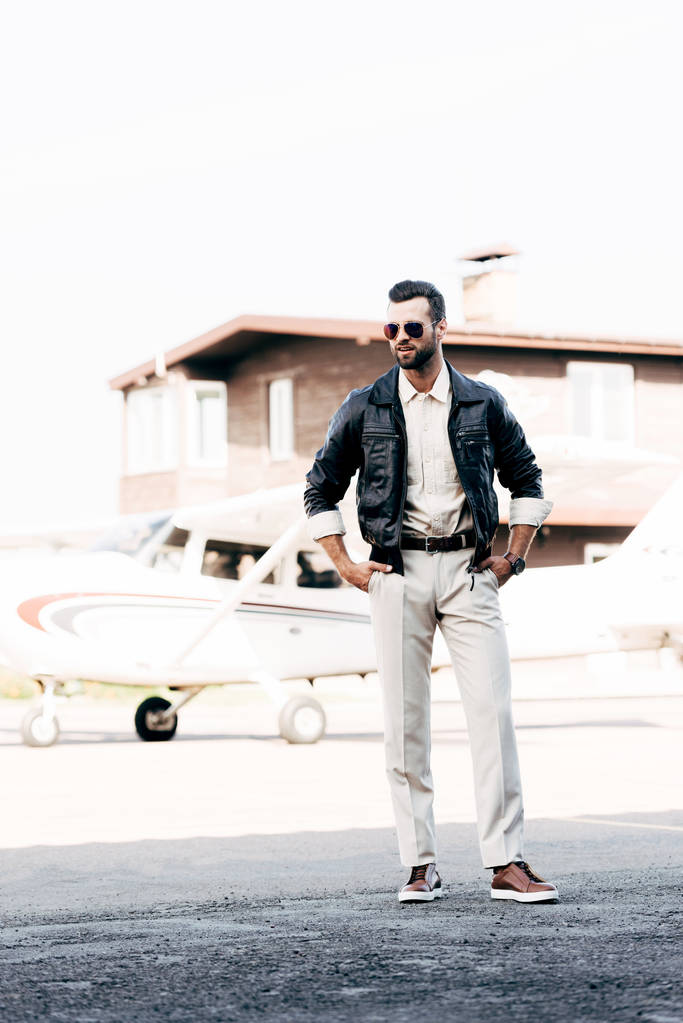 sonriente piloto masculino de moda en chaqueta de cuero y gafas de sol de pie con las manos en la cintura cerca del avión
 - Foto, imagen