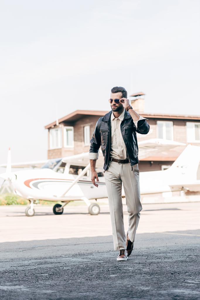 handsome stylish man in leather jacket and sunglasses walking near airplane  - Photo, Image