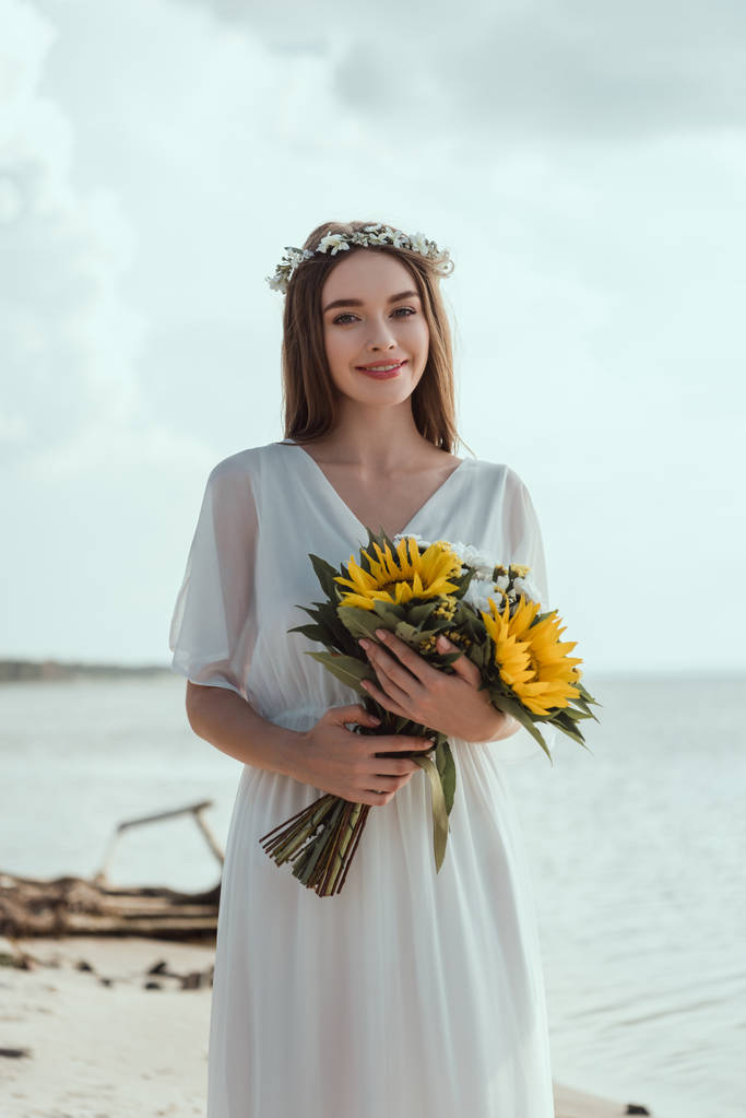 junge Frau in elegantem weißen Kleid und Blumenkranz mit Sonnenblumen am Meeresufer - Foto, Bild