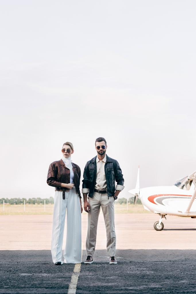 jovem casal na moda em óculos de sol e jaquetas de couro de pé perto do avião
 - Foto, Imagem