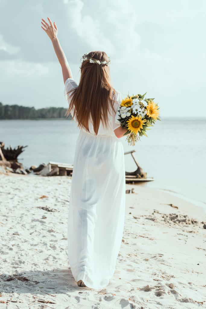 Вид сзади девушки в белом платье, держащей букет с подсолнухами на берегу
 - Фото, изображение