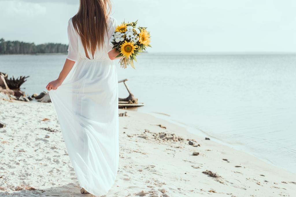 Rückansicht einer Frau im weißen Kleid, die einen Strauß mit Sonnenblumen am Strand hält - Foto, Bild