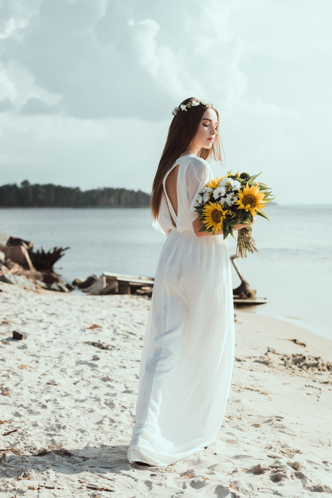 елегантна дівчина в білій сукні і квітковому вінку тримає квіти на морському березі
 - Фото, зображення