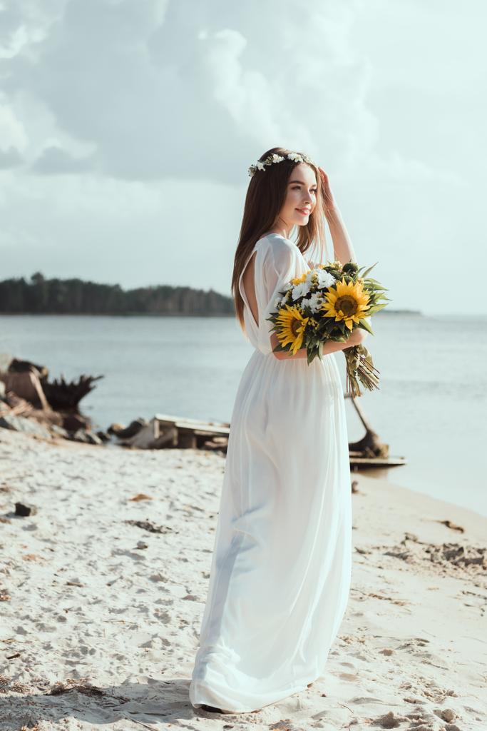 attractive girl in elegant dress and floral wreath holding sunflowers on seashore - Photo, Image