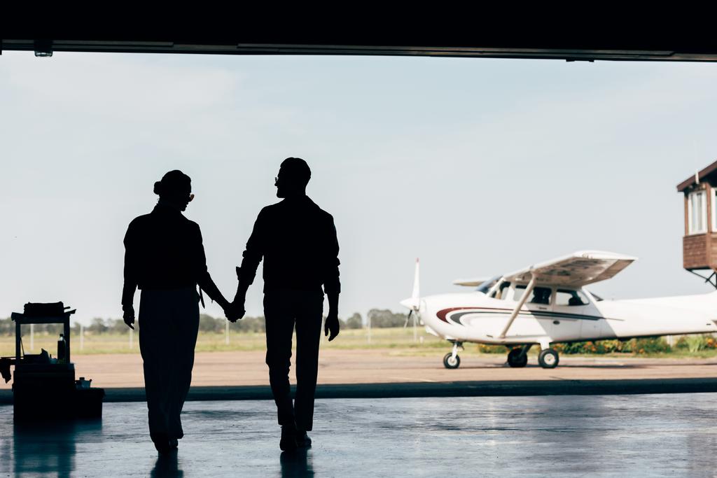 Achteraanzicht van de silhouetten van jonge paar hand in hand en wandelen in de hangar in de buurt van vliegtuig  - Foto, afbeelding