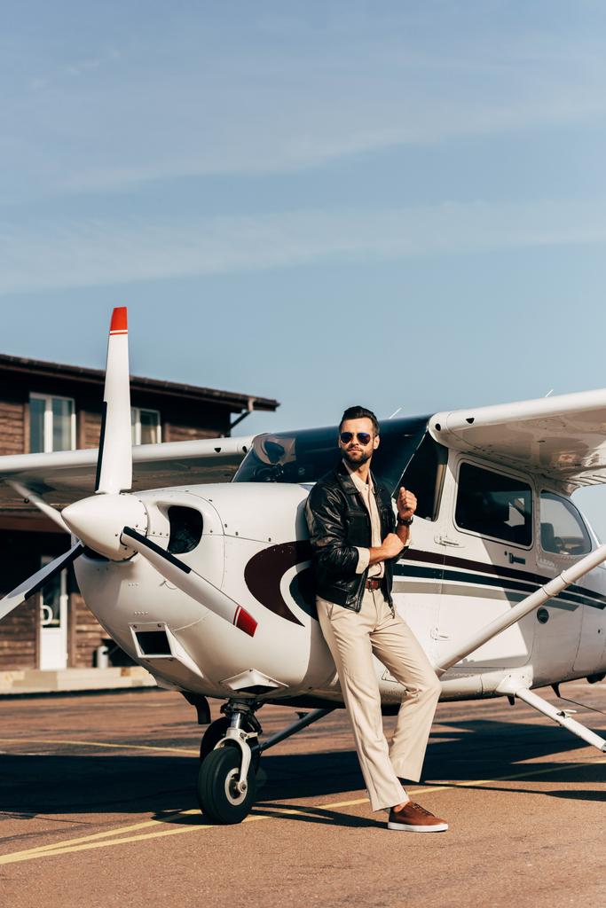 κομψό άνδρα σε δερμάτινο μπουφάν και γυαλιά ηλίου που στέκεται κοντά στο αεροπλάνο  - Φωτογραφία, εικόνα