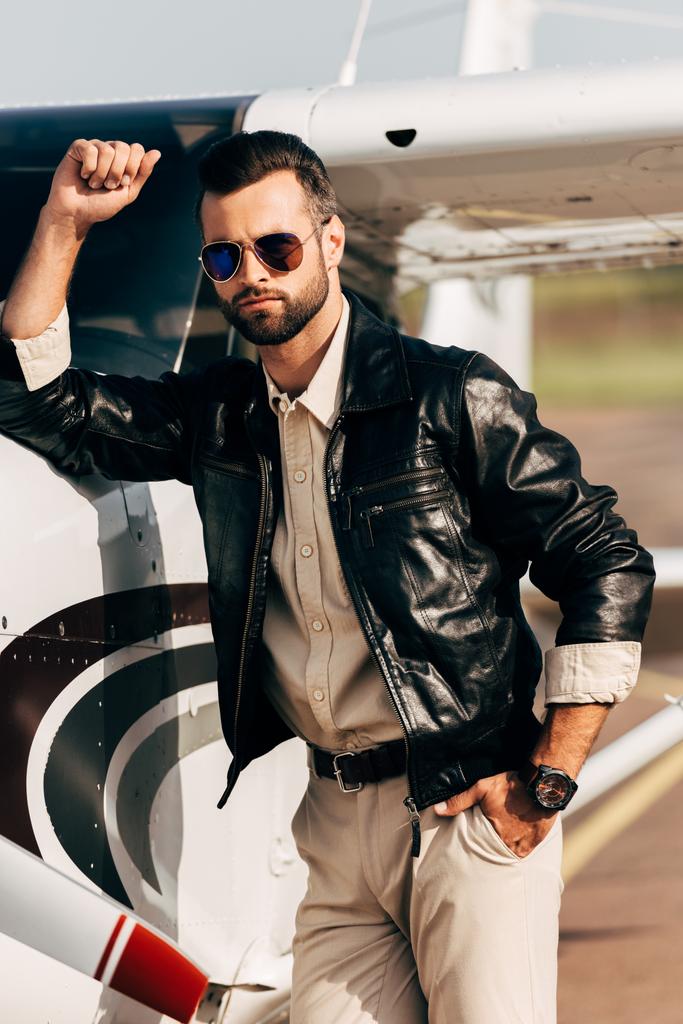 серйозний модний чоловічий пілот у шкіряній куртці та сонцезахисних окулярах, що стоять біля літака
 - Фото, зображення