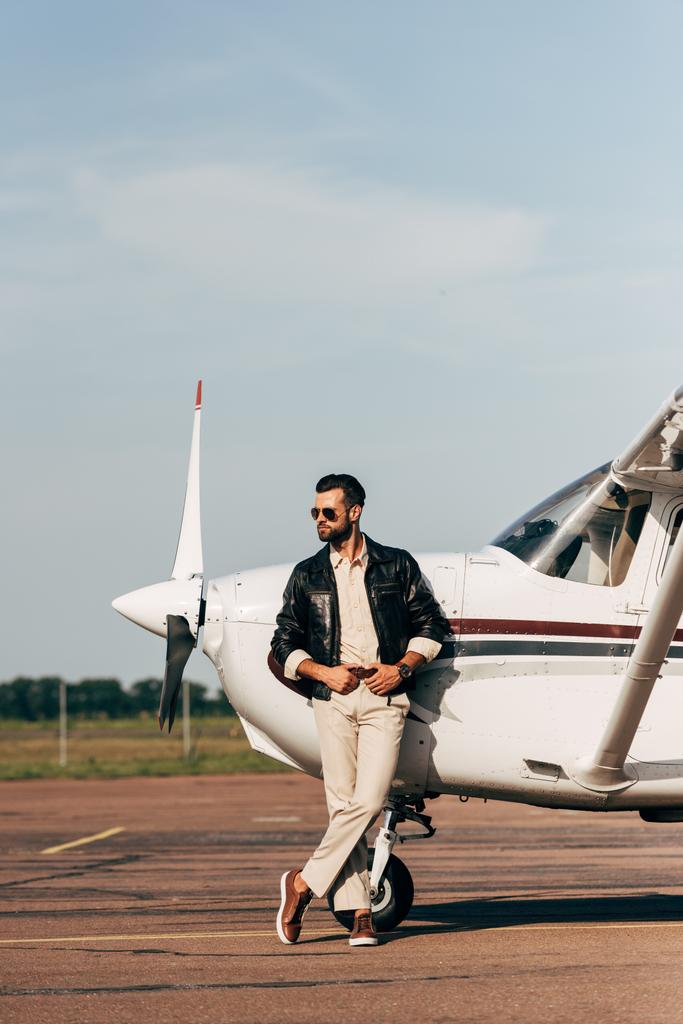hombre guapo en chaqueta de cuero y gafas de sol posando cerca de avión
 - Foto, Imagen