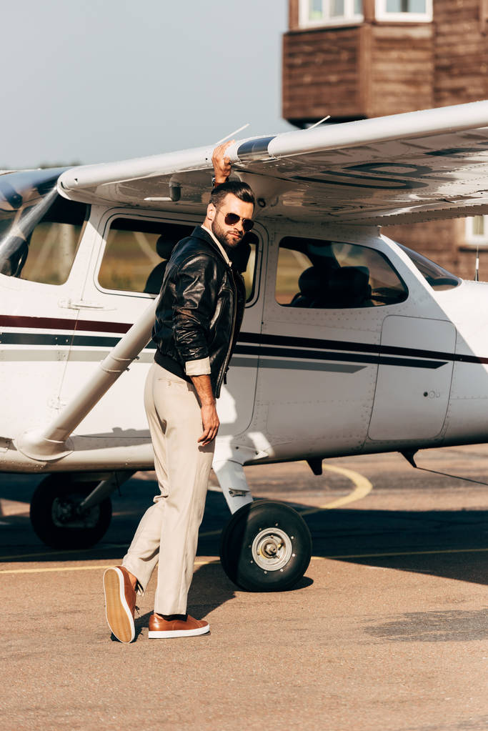 bonito elegante piloto masculino em jaqueta de couro e óculos de sol de pé perto da aeronave
  - Foto, Imagem