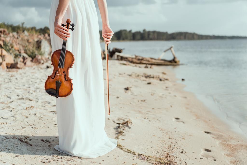 Ausgeschnittene Ansicht eines eleganten Mädchens im weißen Kleid mit Geige am Sandstrand - Foto, Bild