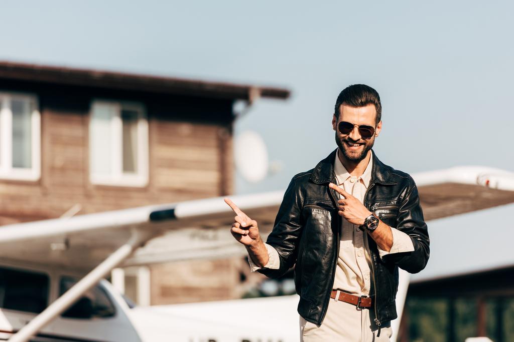 glücklicher stylischer Mann in Lederjacke und Sonnenbrille, der mit den Fingern auf Flugzeug zeigt  - Foto, Bild