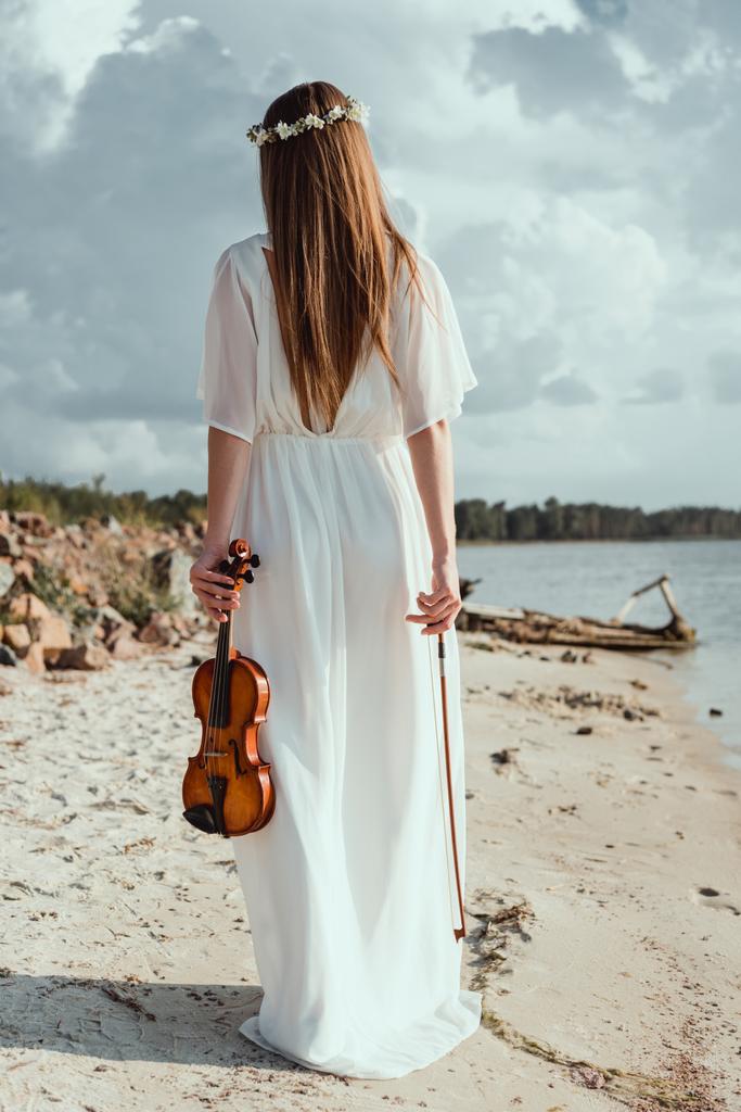 Achteraanzicht van meisje in elegante witte jurk bedrijf viool aan kust - Foto, afbeelding