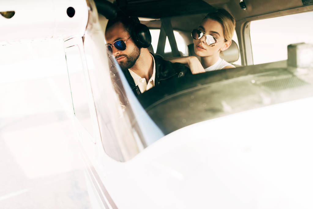 αρσενικό πιλότος στα ακουστικά και γυαλιά ηλίου βλέπει μακριά και να κάθεται με την κοπέλα στην καμπίνα του αεροπλάνου - Φωτογραφία, εικόνα