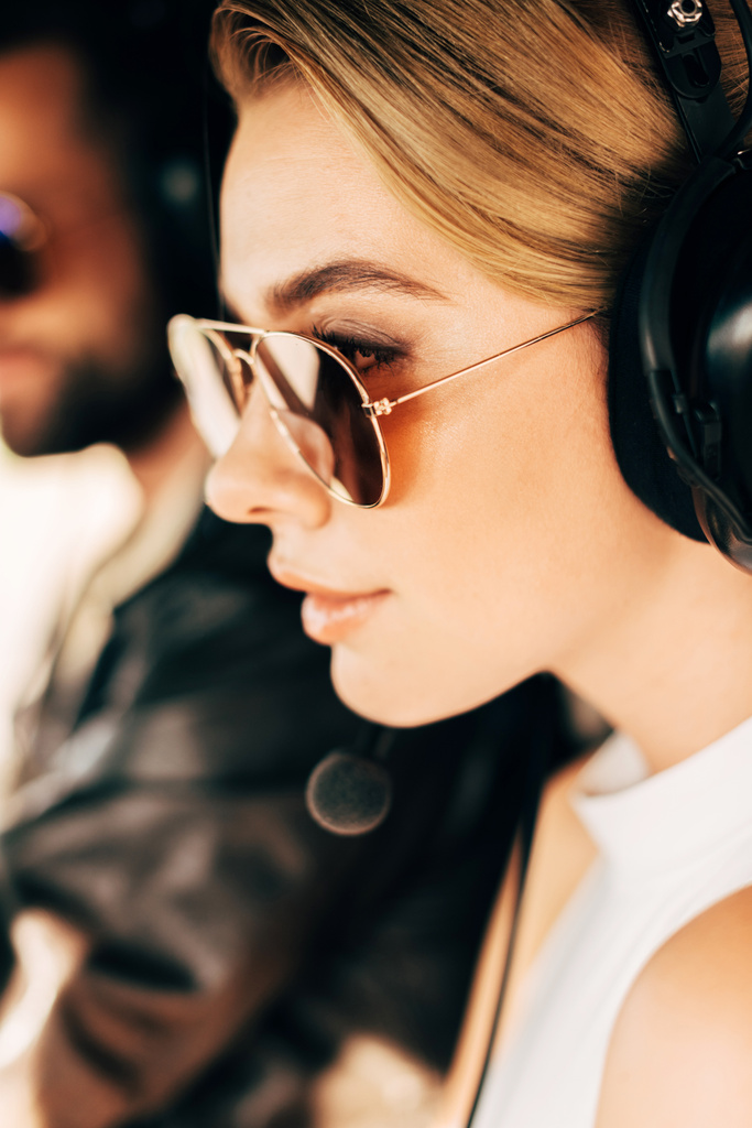 Selektivní fokus krásná žena v sluneční brýle a sluchátka s mikrofonem poblíž mužské pilot v kabině letadla  - Fotografie, Obrázek