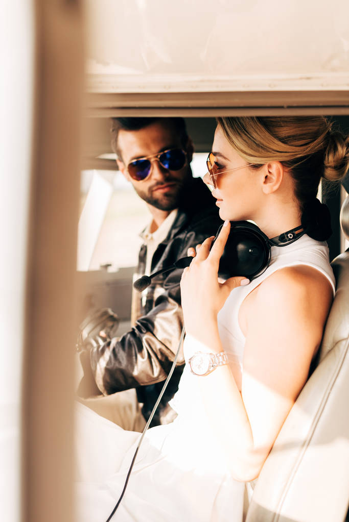 vista lateral de piloto masculino serio en gafas de sol sentado con novia en cabina de avión
 - Foto, Imagen