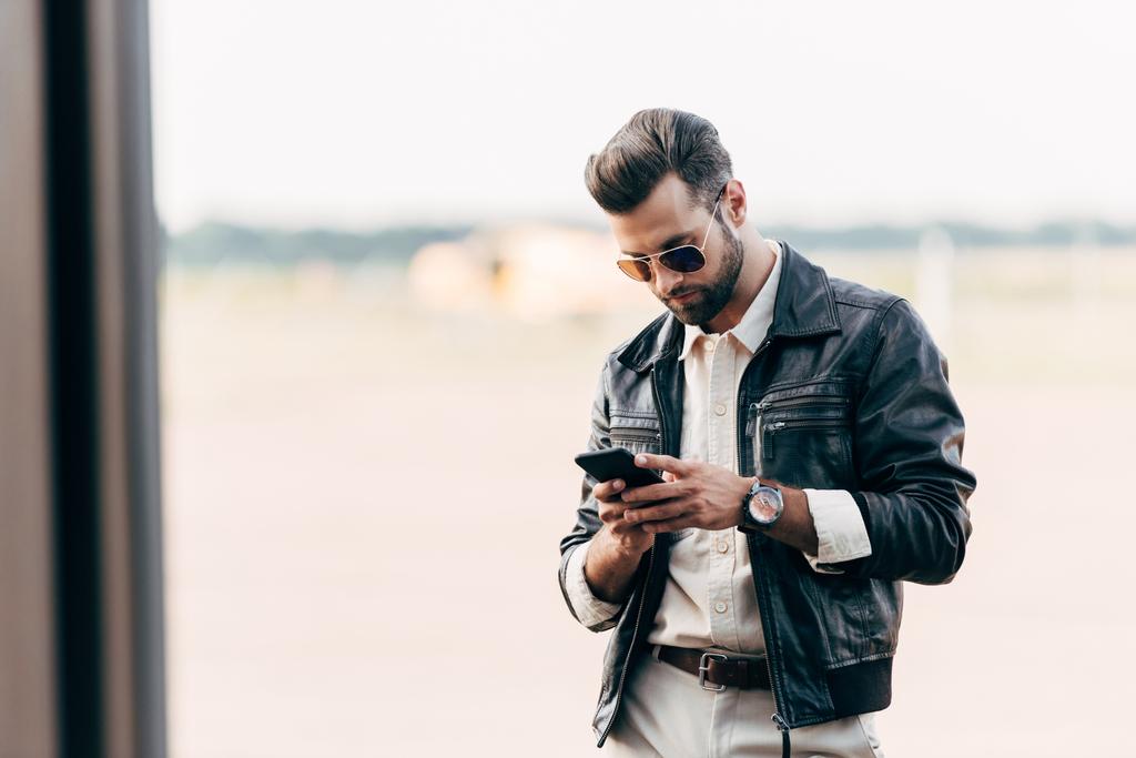 hombre guapo de moda en chaqueta de cuero y gafas de sol usando teléfono inteligente
  - Foto, Imagen