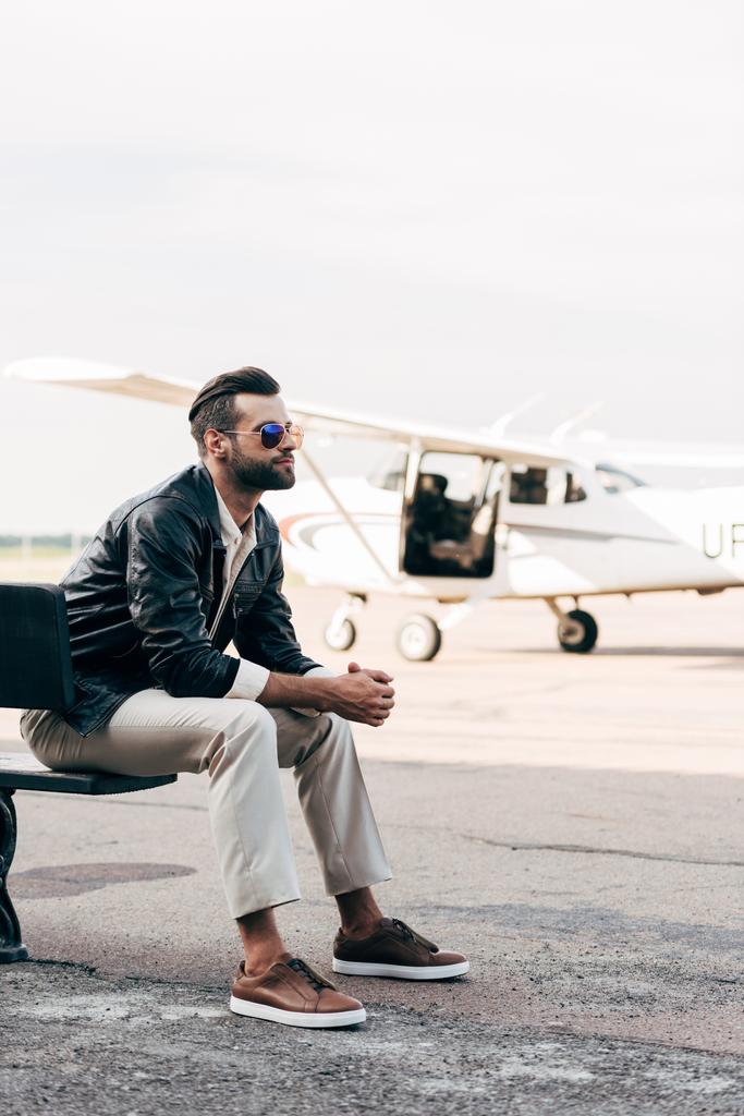 Stilvoller junger männlicher Pilot mit Sonnenbrille sitzt auf Bank in der Nähe des Flugzeugs  - Foto, Bild
